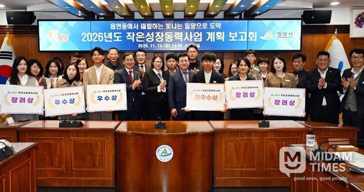 밀양시청 소회의실에서 열린 2026년도 작은성장동력사업 계획 보고회에서, 평가 결과 선정된 읍·면·동 직원들이 사업 선정 표찰을 들고 단체 사진을 촬영하고 있다.